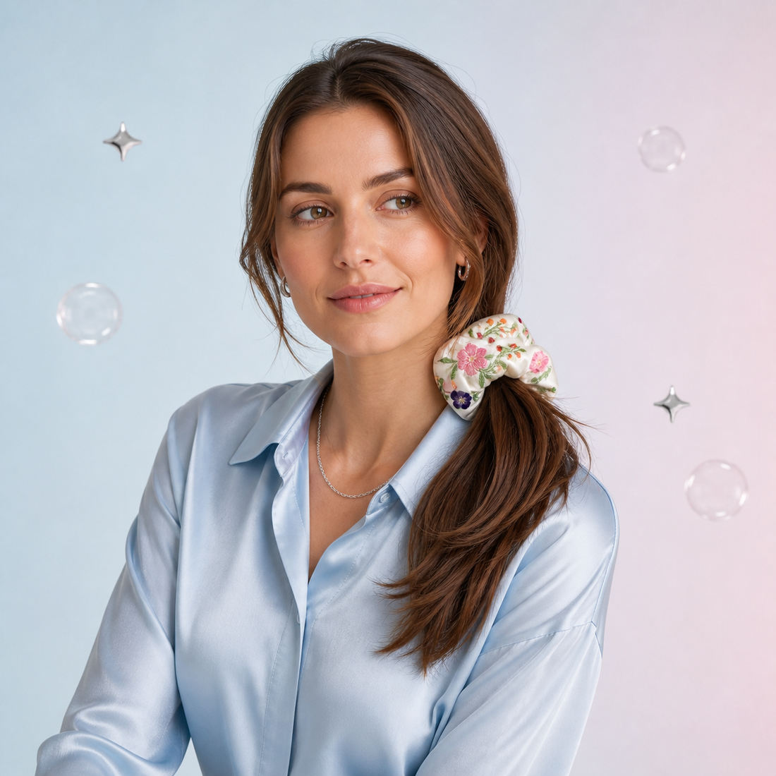 A woman with brown hair wearing a floral-patterned scrunchie, styled in a ponytail, wearing a light blue satin shirt against a pastel pink and blue background with floating bubbles.