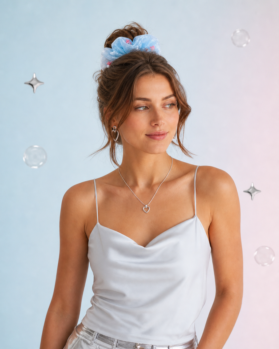 A woman wearing a white camisole top and silver heart necklace, with a light blue scrunchie in her hair, posed against a pastel blue and pink background with floating bubbles and star decorations.