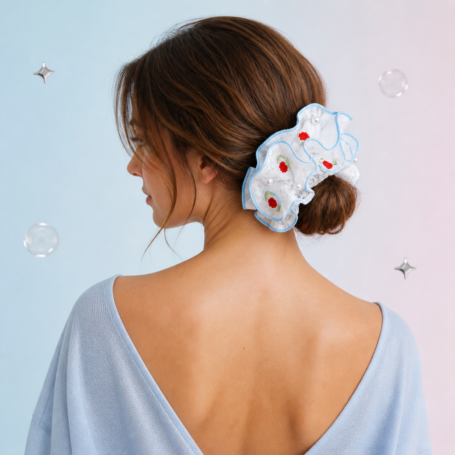 A woman shown from the back wearing a light blue and white ruffled scrunchie with red embroidered details in her brown hair, set against a pastel blue and pink background with floating bubbles.
