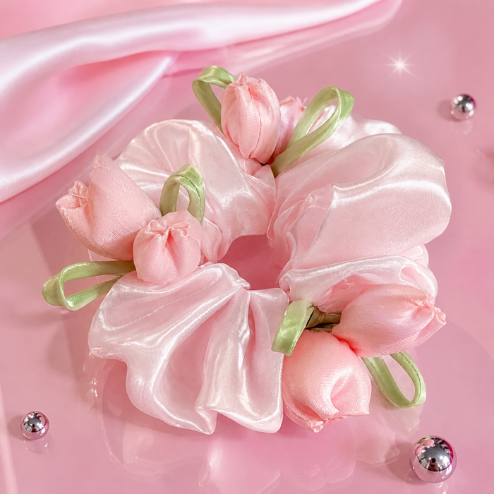 A pink satin hair scrunchie decorated with small pink tulip flowers and light green ribbon accents, photographed on a glossy pink surface with decorative rhinestone-like beads.